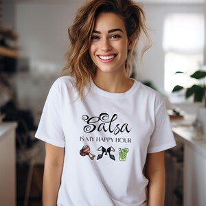 Puede incluir: Una camiseta blanca de manga corta con el texto negro "Salsa IS MY HAPPY HOUR" impreso en el pecho. Debajo del texto hay tres pequeñas ilustraciones coloridas: un par de maracas, un par de zapatos de baile negros y un vaso de cóctel verde.