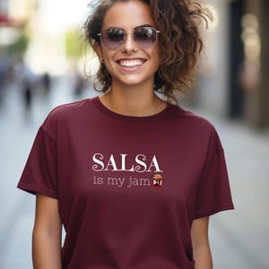 以下が含まれることがあります： 「SALSA is my jam」の白い文字がプリントされた、マルーン色のTシャツ。テキストの下には、小さなサルサジャーのグラフィックがあります。シャツは無地で、クルーネックです。