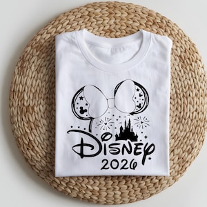 May include: White t-shirt featuring a black graphic of Mickey Mouse ears with stars, a bow, fireworks, and a castle. The word "Disney" is written in script, with "2026" below it. The shirt is folded on a woven, circular mat.
