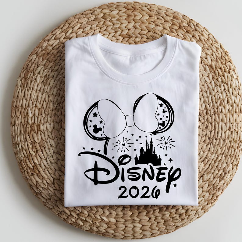 May include: White t-shirt featuring a black graphic of Mickey Mouse ears with stars, a bow, fireworks, and a castle. The word "Disney" is written in script, with "2026" below it. The shirt is folded on a woven, circular mat.