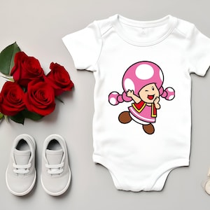 Pode incluir: Body de beb&eacute; branco com uma personagem de desenho animado com um chap&eacute;u de cogumelo rosa com bolinhas brancas. A personagem est&aacute; a piscar o olho e usa um vestido rosa. Rosas vermelhas e sapatos de beb&eacute; cinzentos tamb&eacute;m est&atilde;o na imagem.