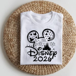 May include: White t-shirt featuring a black Mickey Mouse head design filled with stars. Below the head is the word "Disney" in script, with "2026" underneath. The shirt is displayed on a woven, circular placemat.