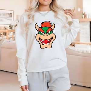 Pode incluir: Camisola branca com um gr&aacute;fico de desenho animado colorido do rosto de Bowser. O gr&aacute;fico inclui cores vermelhas, verdes, laranja e bege. A camisola tem mangas compridas e gola redonda.