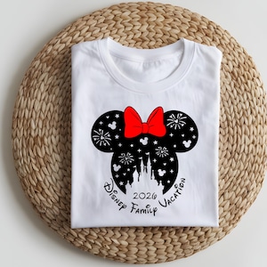 May include: White t-shirt featuring a black Minnie Mouse silhouette with a red bow. The design includes fireworks, a castle, and the text "Disney Family Vacation 2026". The shirt is folded on a woven mat.