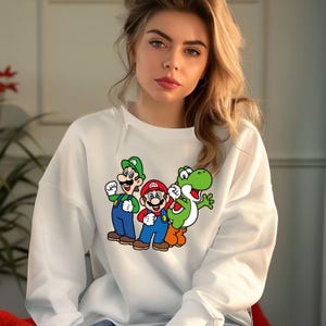 Pode incluir: Camisola branca com um gr&aacute;fico colorido de Mario, Luigi e Yoshi. As personagens de desenhos animados usam seus trajes caracter&iacute;sticos: vermelho, verde, azul e laranja. A camisola &eacute; de estilo casual e confort&aacute;vel.