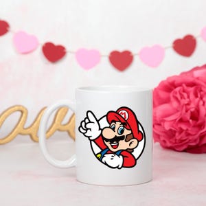 Pode incluir: Caneca de cer&acirc;mica branca com uma ilustra&ccedil;&atilde;o colorida de Mario, o personagem de videogame, apontando para cima. A caneca tem uma al&ccedil;a curva e um design circular. O fundo inclui uma flor rosa e uma guirlanda em forma de cora&ccedil;&atilde;o.