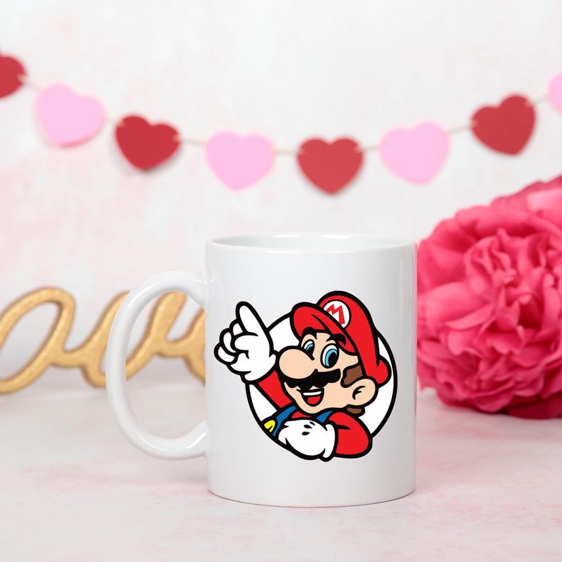 Pode incluir: Caneca de cer&acirc;mica branca com uma ilustra&ccedil;&atilde;o colorida de Mario, o personagem de videogame, apontando para cima. A caneca tem uma al&ccedil;a curva e um design circular. O fundo inclui uma flor rosa e uma guirlanda em forma de cora&ccedil;&atilde;o.