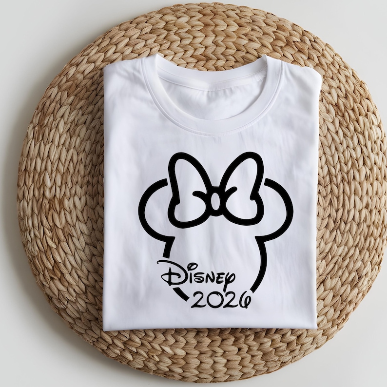May include: White t-shirt featuring a black graphic of Minnie Mouse's head with a bow and the text "Disney 2026". The shirt is folded and placed on a woven, circular mat.