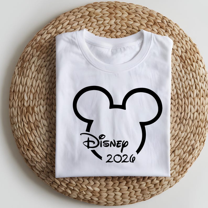 May include: A white t-shirt featuring a black outline of Mickey Mouse's head with the word "Disney" and the year "2026" printed in black. The shirt is folded and placed on a woven, circular mat.