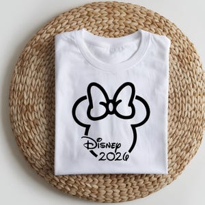 May include: White t-shirt featuring a black Minnie Mouse silhouette with a bow, the word "Disney" in script, and the year "2026". The shirt is folded neatly on a woven, circular placemat.