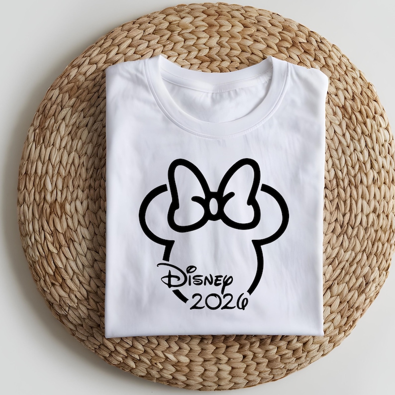 May include: White t-shirt featuring a black Minnie Mouse silhouette with a bow, the word "Disney" in script, and the year "2026". The shirt is folded neatly on a woven, circular placemat.