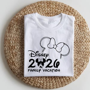 May include: White t-shirt with a black graphic design featuring the Disney logo, the year "2020", and the words "Family Vacation". The design includes a Minnie Mouse bow and ears.
