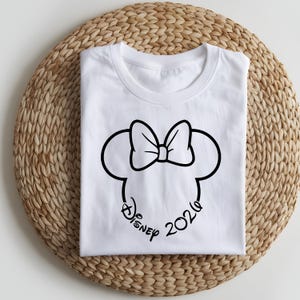 May include: White t-shirt featuring a black outline of a bow and mouse ears, with the text "Disney 2020" in a script font. The shirt is folded neatly on a woven, circular placemat.