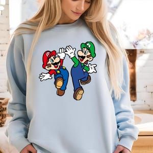 Pode incluir: Camisola azul clara com um desenho de Mario e Luigi a cumprimentarem-se. Mario usa um chap&eacute;u vermelho, camisa vermelha e macac&atilde;o azul. Luigi usa um chap&eacute;u verde, camisa verde e macac&atilde;o azul. Ambos personagens usam luvas brancas e sapatos castanhos.