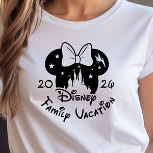May include: White t-shirt featuring a black graphic of a mouse head with a bow, stars, and a castle silhouette. The text reads "20 Disney Family Vacation 26". A fun shirt for a family trip.