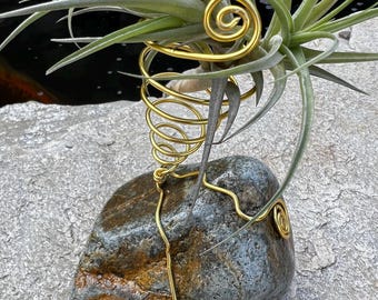 May include: A decorative air plant holder featuring a natural stone base and a gold-colored wire design. The wire is shaped into spirals and curves, cradling the air plant with its green leaves. The holder sits on a gray stone surface.