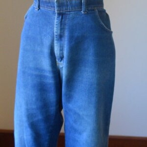 May include: Blue denim jeans with a high waist and flared leg, displayed on a mannequin. The jeans have a classic five-pocket design and a button and zip closure. The fabric has a faded appearance.