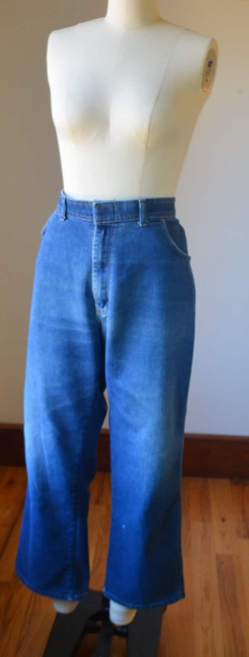 May include: Blue denim jeans with a high waist and flared leg, displayed on a mannequin. The jeans have a classic five-pocket design and a button and zip closure. The fabric has a faded appearance.