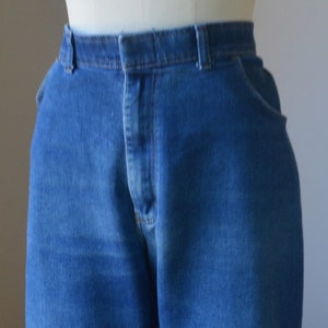May include: Close-up of blue denim jeans on a mannequin. The jeans feature a classic five-pocket design, a button and zip closure, and belt loops. The denim fabric has a medium wash and a slightly faded appearance.