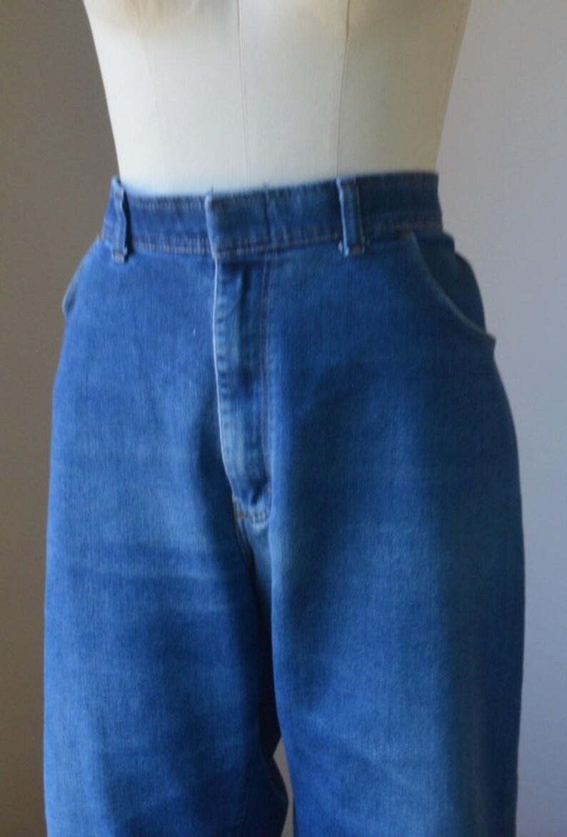 May include: Close-up of blue denim jeans on a mannequin. The jeans feature a classic five-pocket design, a button and zip closure, and belt loops. The denim fabric has a medium wash and a slightly faded appearance.