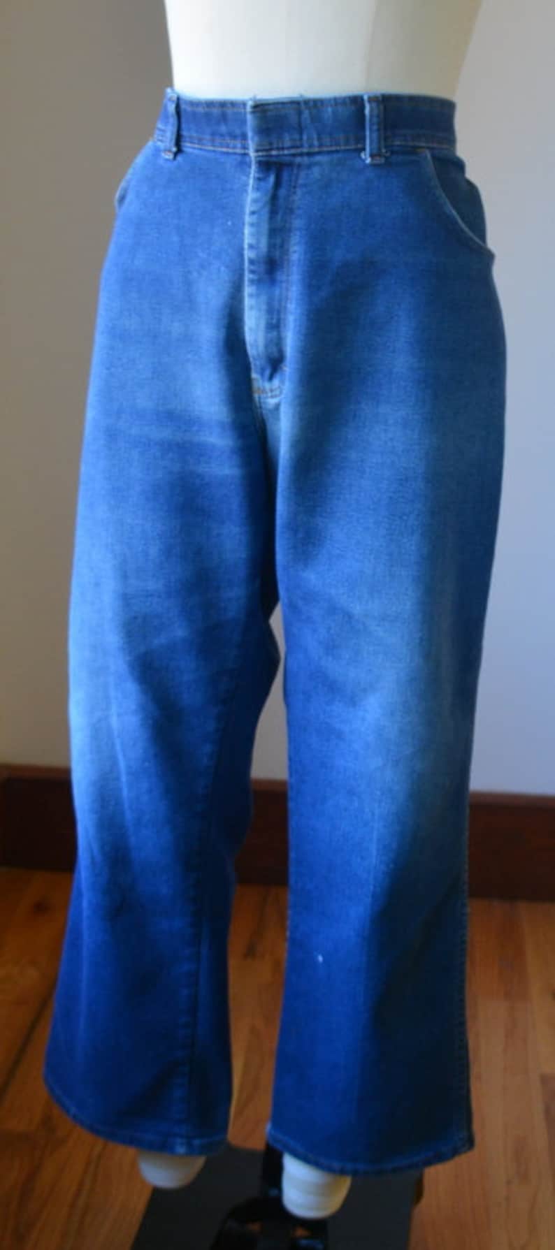 May include: Blue denim jeans with a high waist and flared legs. The jeans are displayed on a mannequin, showcasing the classic design and fit. The fabric has a slight fade, adding to the vintage aesthetic.