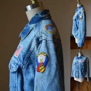 Può includere: Giacca di jeans azzurra con diverse toppe. Le toppe includono una bandiera americana, una toppa circolare con una scena di montagna e una toppa con il testo "504 Devils" e "Airborne". La giacca è esposta su un manichino.