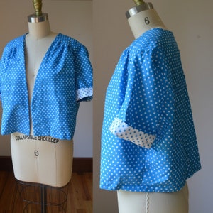 May include: A blue bolero jacket with white polka dots. The short-sleeved jacket has a V-neck and white cuffs. The garment is displayed on a mannequin.