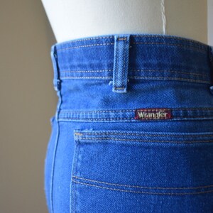 May include: Close-up of the rear of blue denim jeans, featuring the Wrangler logo on the back pocket. The jeans have a classic design with visible stitching and a red logo tag.