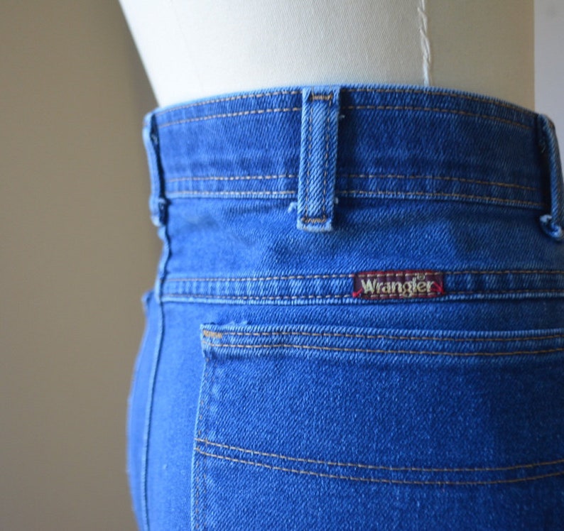 May include: Close-up of the rear of blue denim jeans, featuring the Wrangler logo on the back pocket. The jeans have a classic design with visible stitching and a red logo tag.
