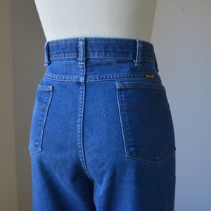 May include: Close-up of the rear of blue denim jeans. The jeans have a high waist, two back pockets, and a Wrangler label. The stitching is visible, and the jeans are displayed on a mannequin.