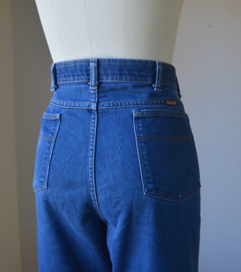 May include: Close-up of the rear of blue denim jeans. The jeans have a high waist, two back pockets, and a Wrangler label. The stitching is visible, and the jeans are displayed on a mannequin.
