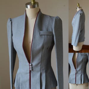 May include: A gray blazer with a tailored fit, featuring a high collar and a contrasting burgundy trim along the edges. The jacket has a small pocket and three decorative buttons. The design includes pleated details at the waist and padded shoulders.