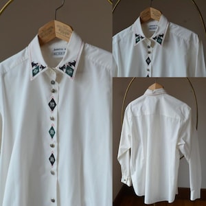 May include: A white long-sleeved shirt with decorative embroidery on the collar and button placket. The embroidery includes green, black, and red details. The shirt is displayed on a wooden hanger. The brand name "Dorothy M. Koret" is visible.
