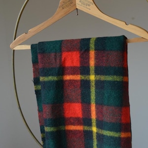 Puede incluir: Una manta a cuadros doblada en tonos verdes, rojos, amarillos y naranjas cuelga de una percha de madera. El patrón de la manta presenta un diseño de tartán clásico. La percha está suspendida de un soporte metálico dorado. La manta mide unos 150 cm de largo.