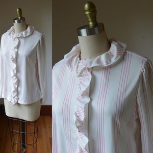 Peut inclure: Un chemisier crème à rayures roses verticales, avec un col et une patte de boutonnage à volants. Le haut à manches longues est présenté sur un mannequin, soulignant son style et son design vintage.