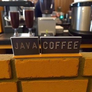 Può includere: Un cartello con la scritta "JAVA COFFEE" in lettere argentate. Il cartello è su un muro di mattoni. Sullo sfondo, c'è una macchina per il caffè, due bicchieri di caffè e una persona.
