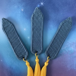 Puede incluir: Tres marcadores de ganchillo hechos a mano. Cada marcador es de color azul apagado con una parte superior puntiaguda y una borla amarilla. Los marcadores están dispuestos sobre un fondo azul y morado con acentos de estrellas blancas.