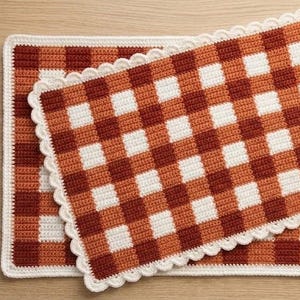 Può includere: Due tovagliette rettangolari all'uncinetto con un motivo a scacchi in tonalità alternate di arancione bruciato, rosso scuro e bianco. Ogni tovaglietta è rifinita con un bordo decorativo smerlato bianco lungo i lati, creando un aspetto materico.