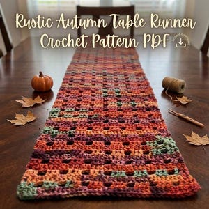 May include: A rectangular crochet table runner featuring a variegated yarn pattern in shades of orange, red, purple, and green. The text "Rustic Autumn Table Runner Crochet Pattern PDF" is displayed above the runner, accompanied by a small pumpkin, dried maple leaves, a spool of twine, and a wooden crochet hook.