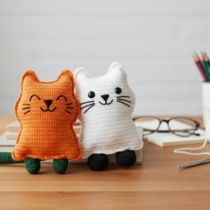 Könnte beinhalten: Zwei handgefertigte Häkeltier-Katzen. Eine ist orange mit grünem Schwanz und Füßen, die andere ist weiß mit schwarzen Füßen. Beide haben gestickte Gesichter mit schwarzen Schnurrhaaren und lächelnden Mäulern. Sie sitzen auf einer Holzoberfläche.