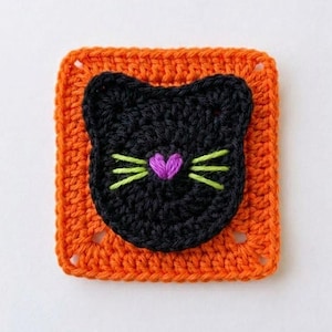 May include: A square crochet granny square featuring a vibrant orange border and a centered black cat face motif. The cat face is detailed with a small purple heart-shaped nose and three lime green embroidered whiskers on each side of the face.