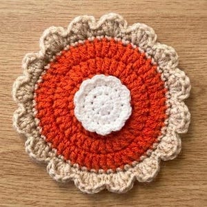 May include: A circular crochet coaster featuring a central white flower-like motif, surrounded by a vibrant orange circular section, and finished with a scalloped beige border. The textile piece displays a layered, textured design with distinct colour zones.