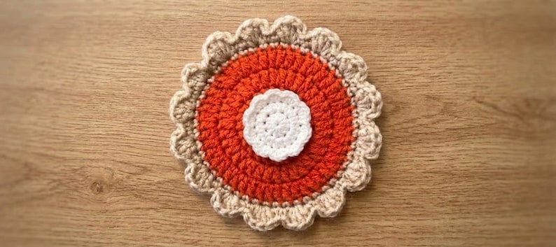 May include: A circular crochet coaster featuring a central white flower-like motif, surrounded by a vibrant orange circular section, and finished with a scalloped beige border. The textile piece displays a layered, textured design with distinct colour zones.