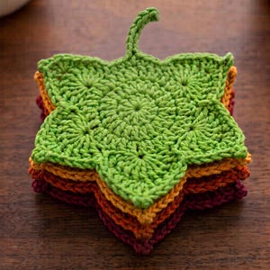 May include: A stack of five crocheted leaf-shaped coasters in a gradient of colors, starting with a bright green top leaf, followed by yellow, orange, red, and deep burgundy layers, each featuring a central stem and textured stitch pattern.