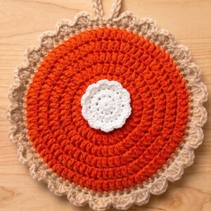 May include: A round, crocheted pot holder designed to resemble a pumpkin pie. The centre is bright orange with a textured, circular stitch pattern, topped with a small, white, crocheted dollop. It features a scalloped, tan-coloured crust edge and a loop.