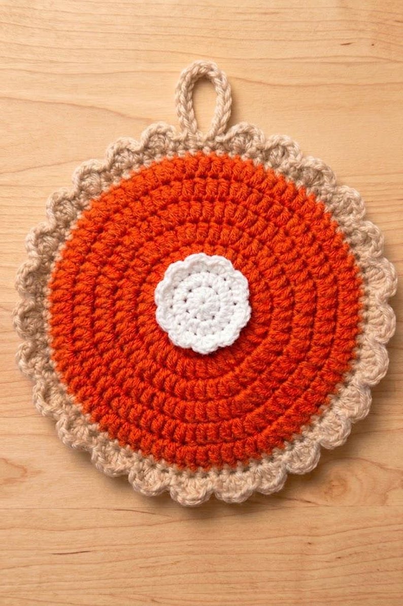 May include: A round, crocheted pot holder designed to resemble a pumpkin pie. The centre is bright orange with a textured, circular stitch pattern, topped with a small, white, crocheted dollop. It features a scalloped, tan-coloured crust edge and a loop.