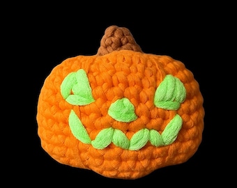 May include: A handcrafted orange crochet pumpkin featuring a brown stem and a stitched jack-o'-lantern face with bright lime green yarn accents for the eyes, nose, and a wide, jagged, toothy grin, all worked in a textured, chunky yarn stitch pattern.