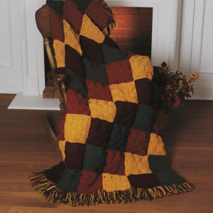 May include: A handmade, checkered throw blanket in shades of gold, burgundy, and forest green. The blanket is draped over a wooden rocking chair, with a fringed edge. The blanket is made of a thick, textured yarn.