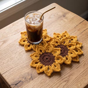 May include: Two sunflower-shaped crochet coasters featuring a dark brown circular centre and bright yellow petals. A clear glass filled with iced coffee and a gold-coloured metal straw sits on top of the coasters, showcasing the intricate yarn work.