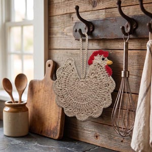 May include: A beige crochet potholder shaped like a chicken with a red comb and wattle and a yellow beak. The potholder features a circular body with scalloped edges and a matching beige loop at the top for hanging on a metal wall hook.